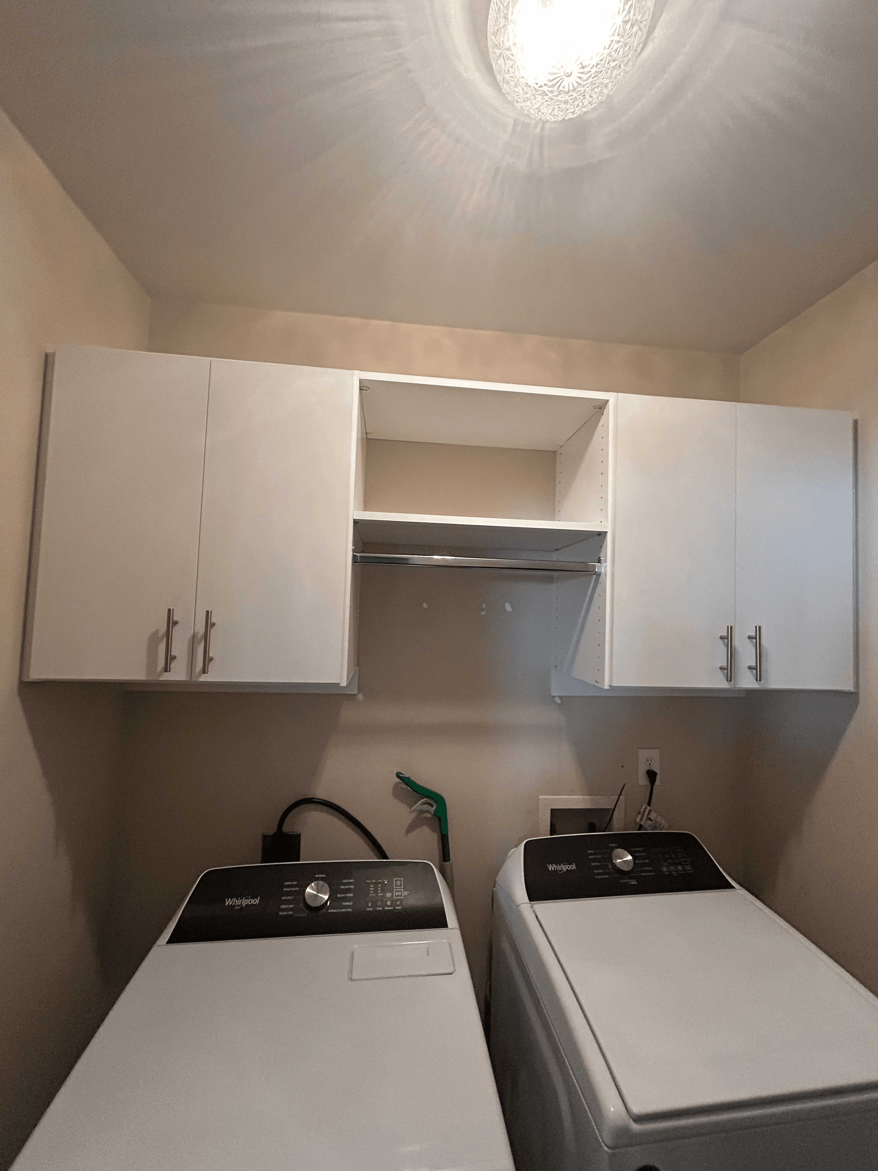 Two white Whirlpool laundry machines below white cabinets with a central open shelf and rod.