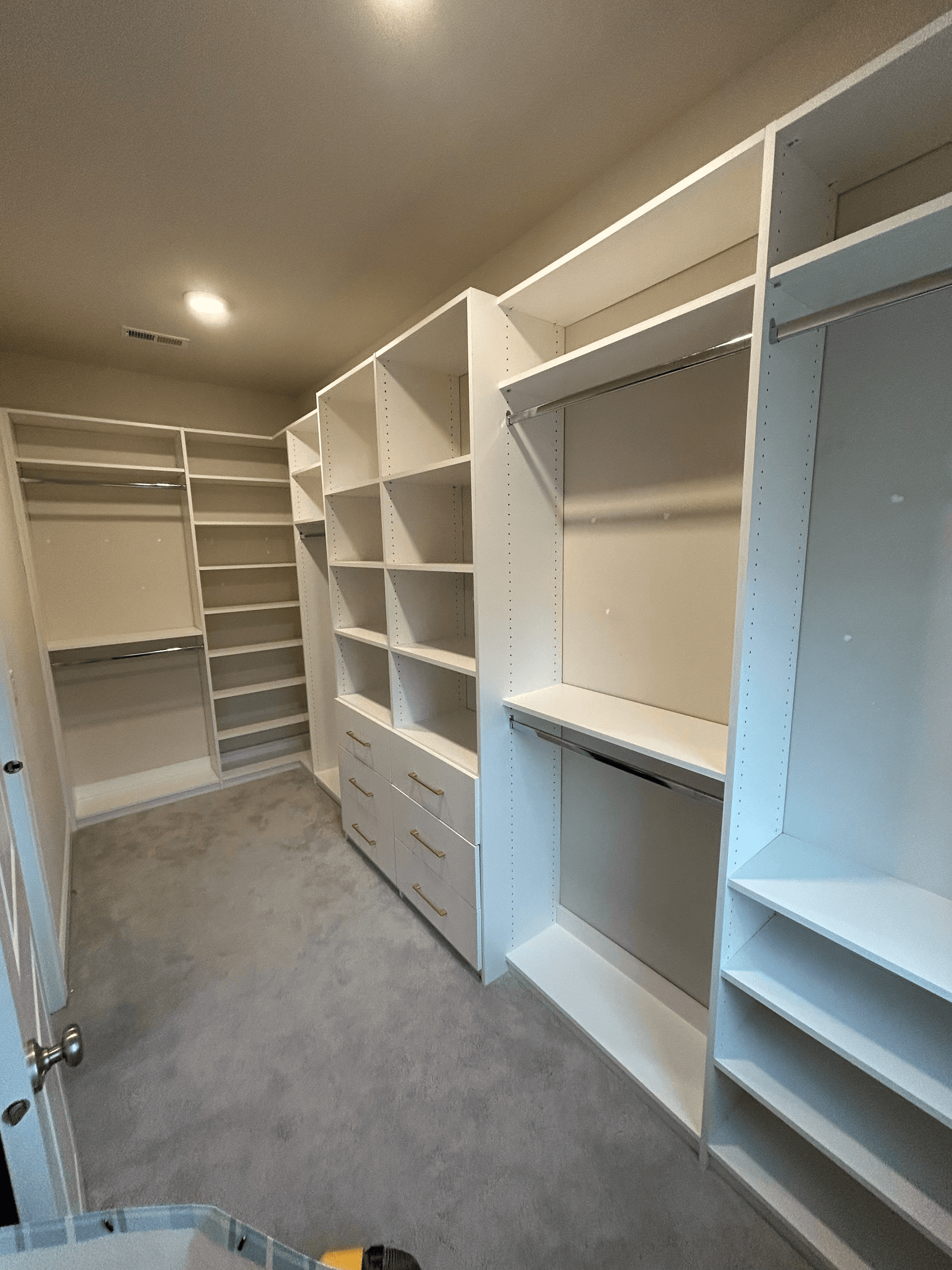 Empty walk-in closet featuring white built-in shelving, drawers, hanging rods, and gray carpeting.