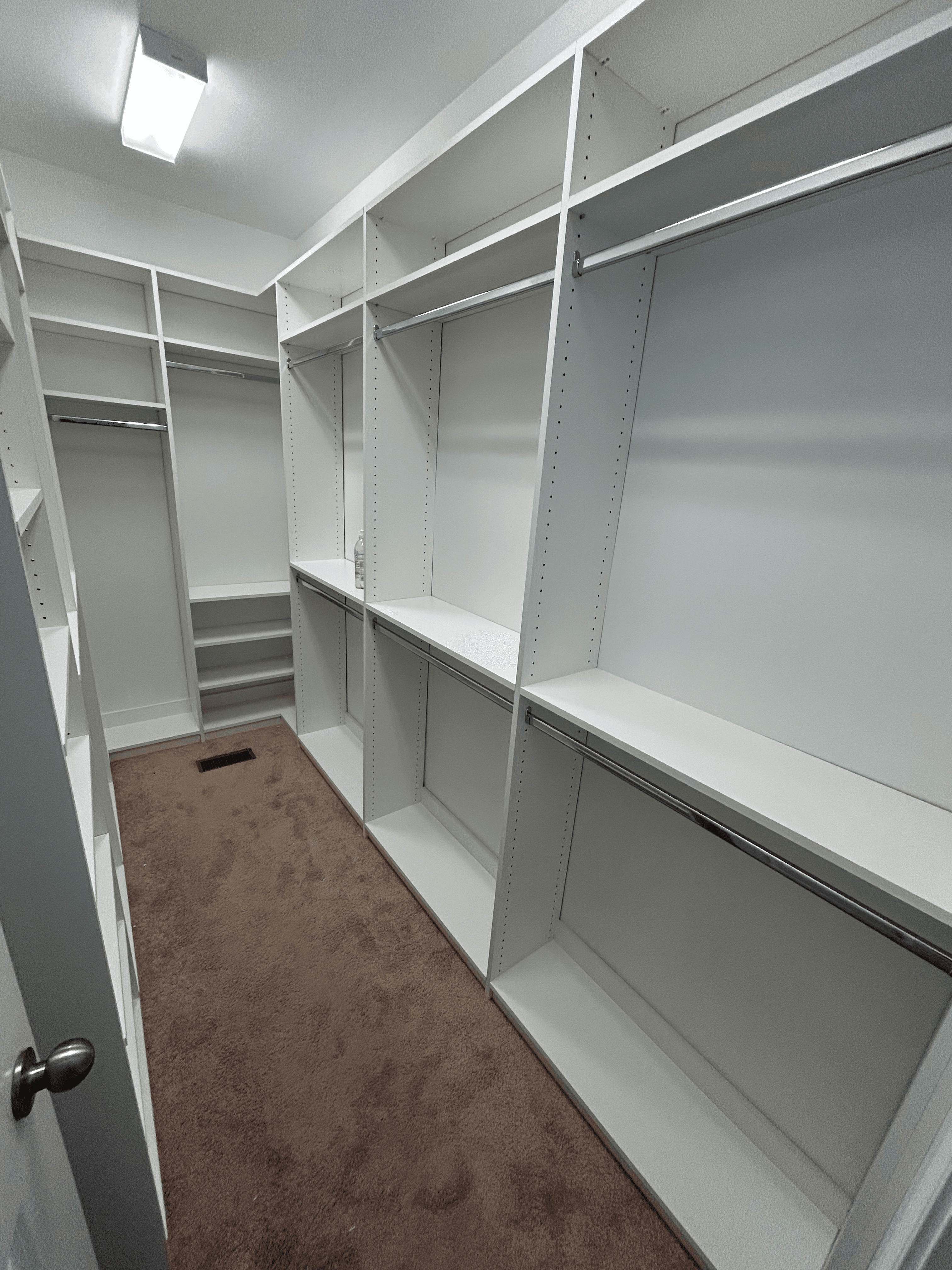 Spacious empty walk-in closet with white built-in shelves, metal hanging rods, and brown carpeting.