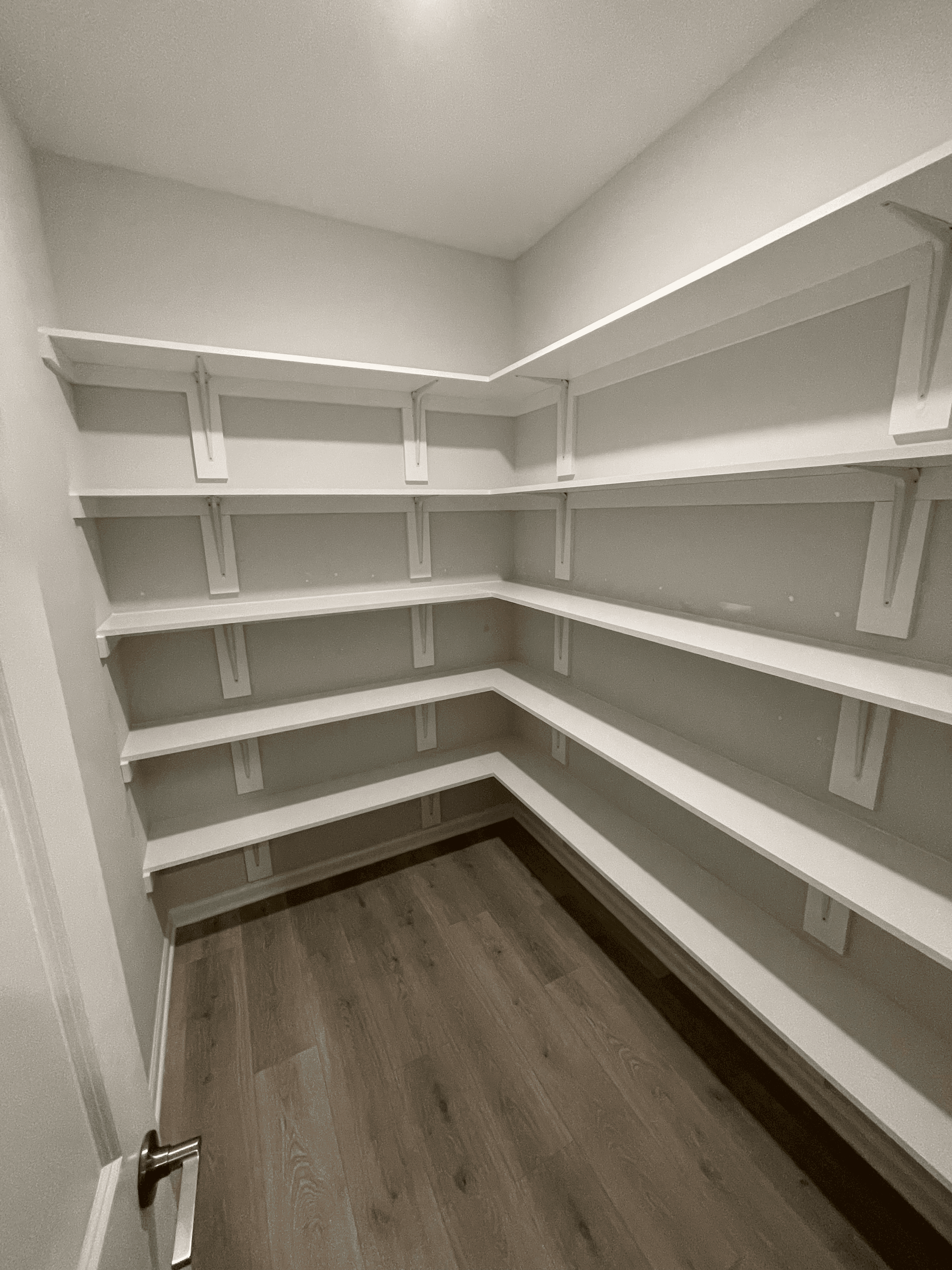Empty white L-shaped shelves wrap around the corner of a pantry with light wood floors.