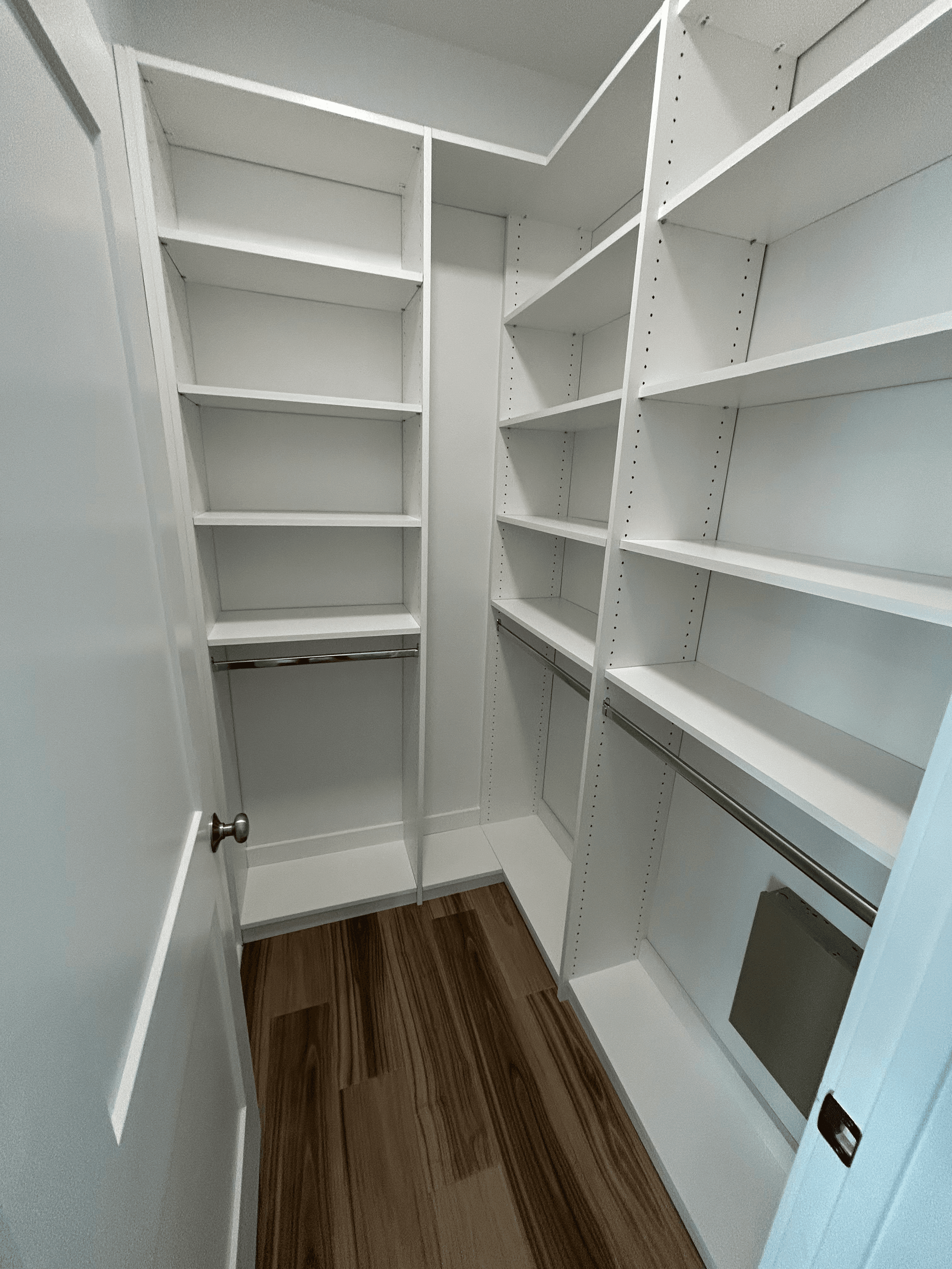 Empty walk-in closet with white built-in shelving, hanging rods, and dark wood laminate flooring.