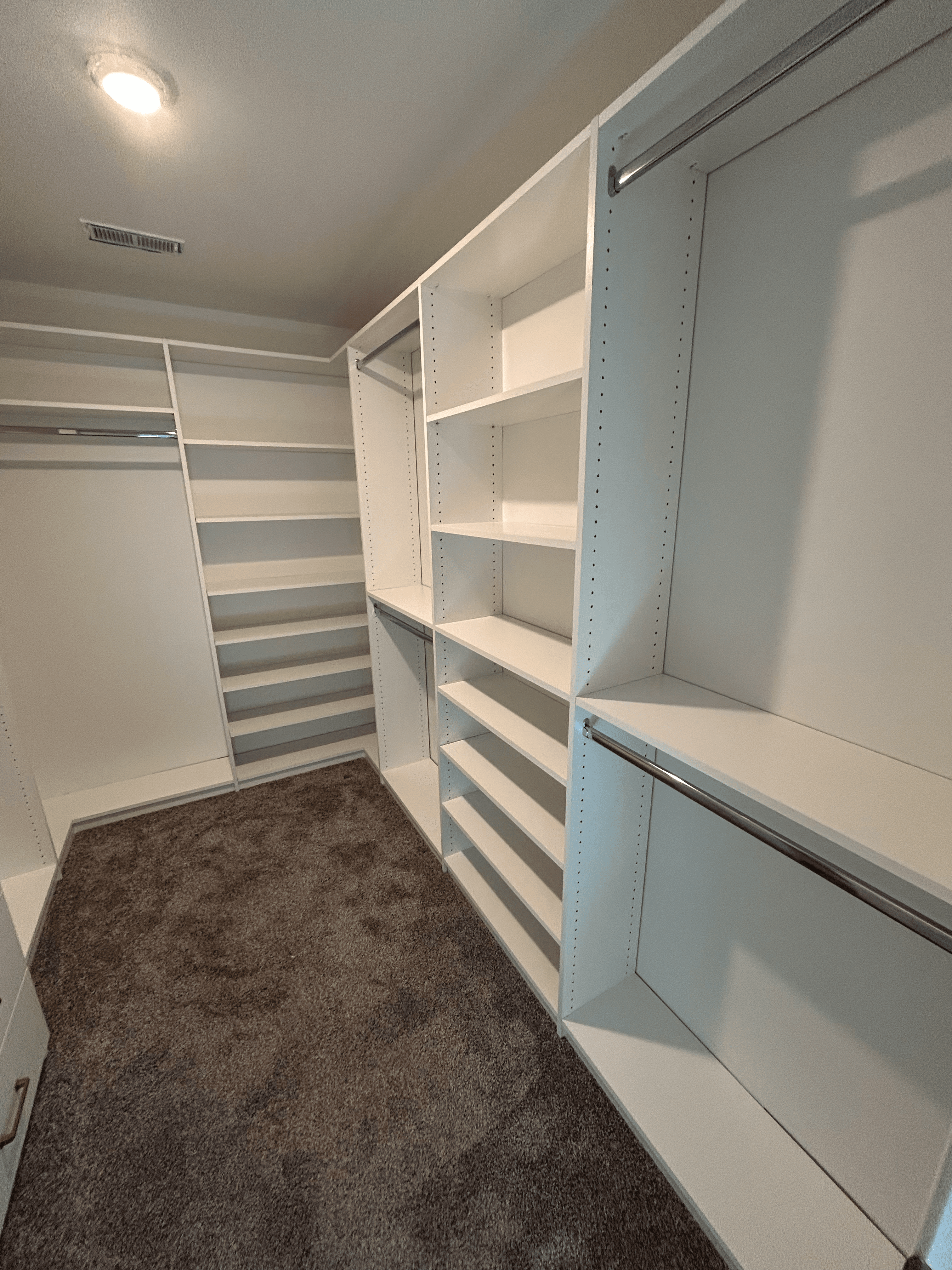 Empty walk-in closet featuring white built-in shelving, metal hanging rods, and dark grey carpeting.