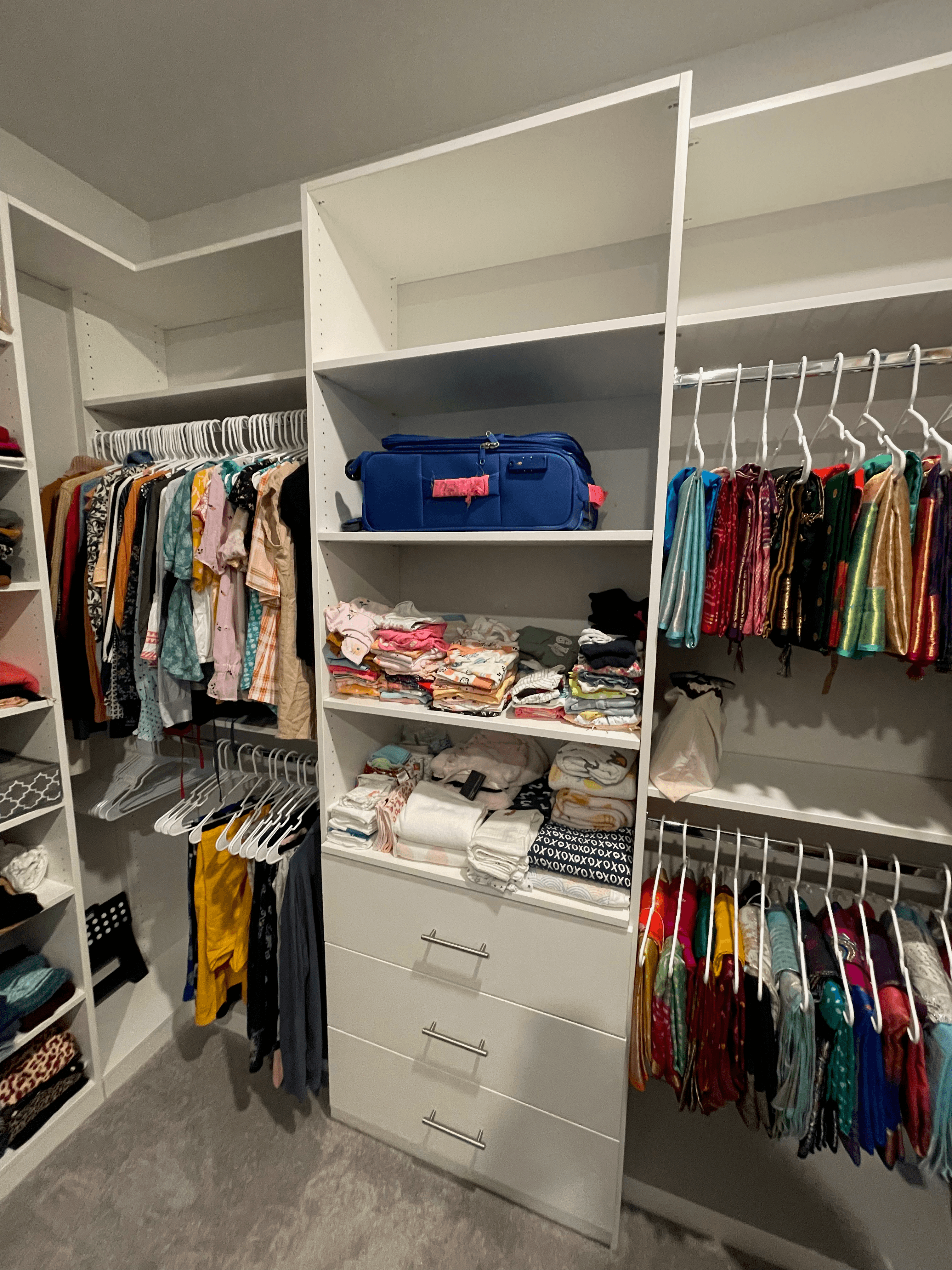 Organized white walk-in closet featuring hanging colorful garments, folded clothes, and a blue suitcase.