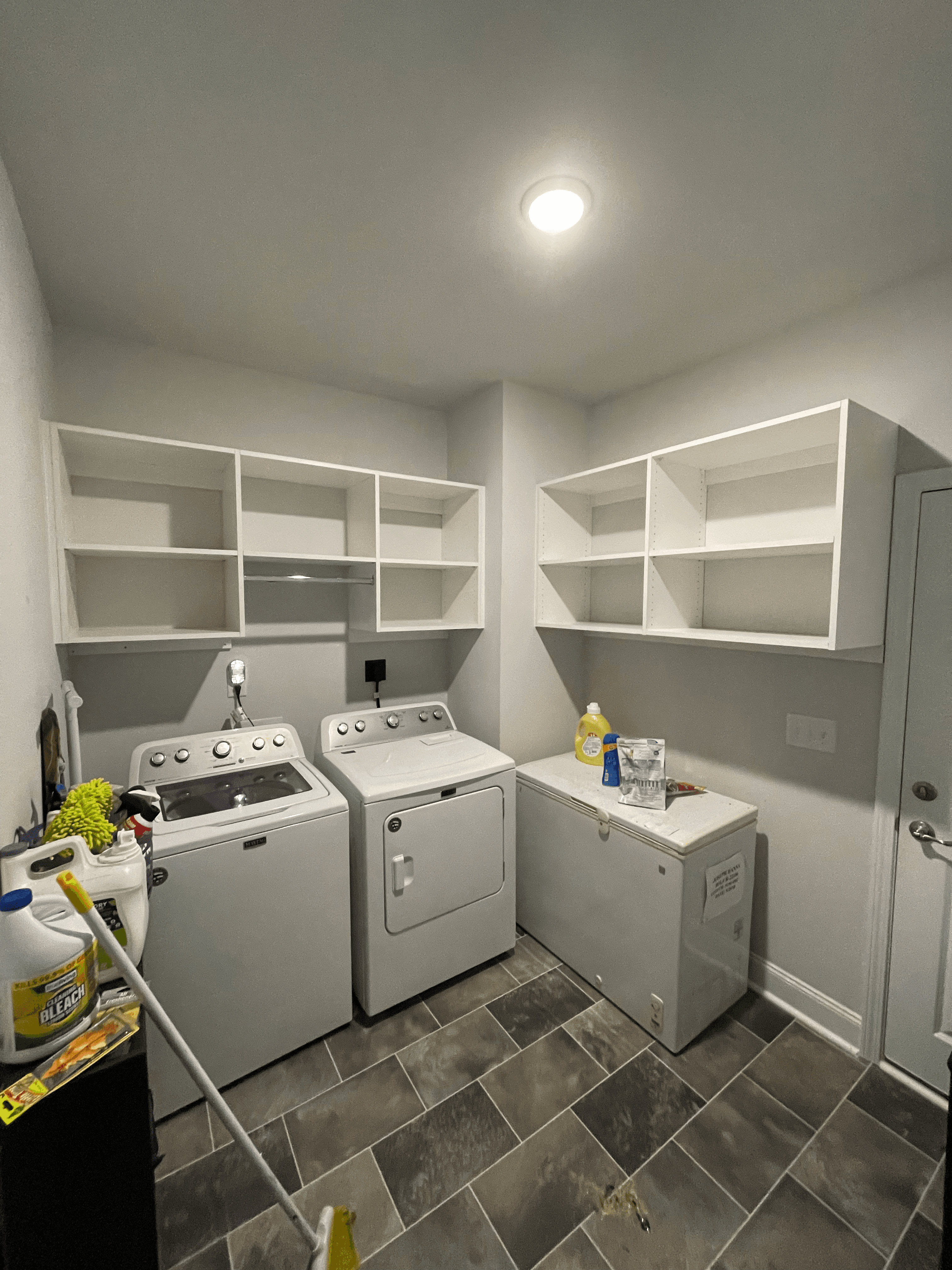 Laundry room with white washer, dryer, chest freezer, and open shelving units on grey tiles.