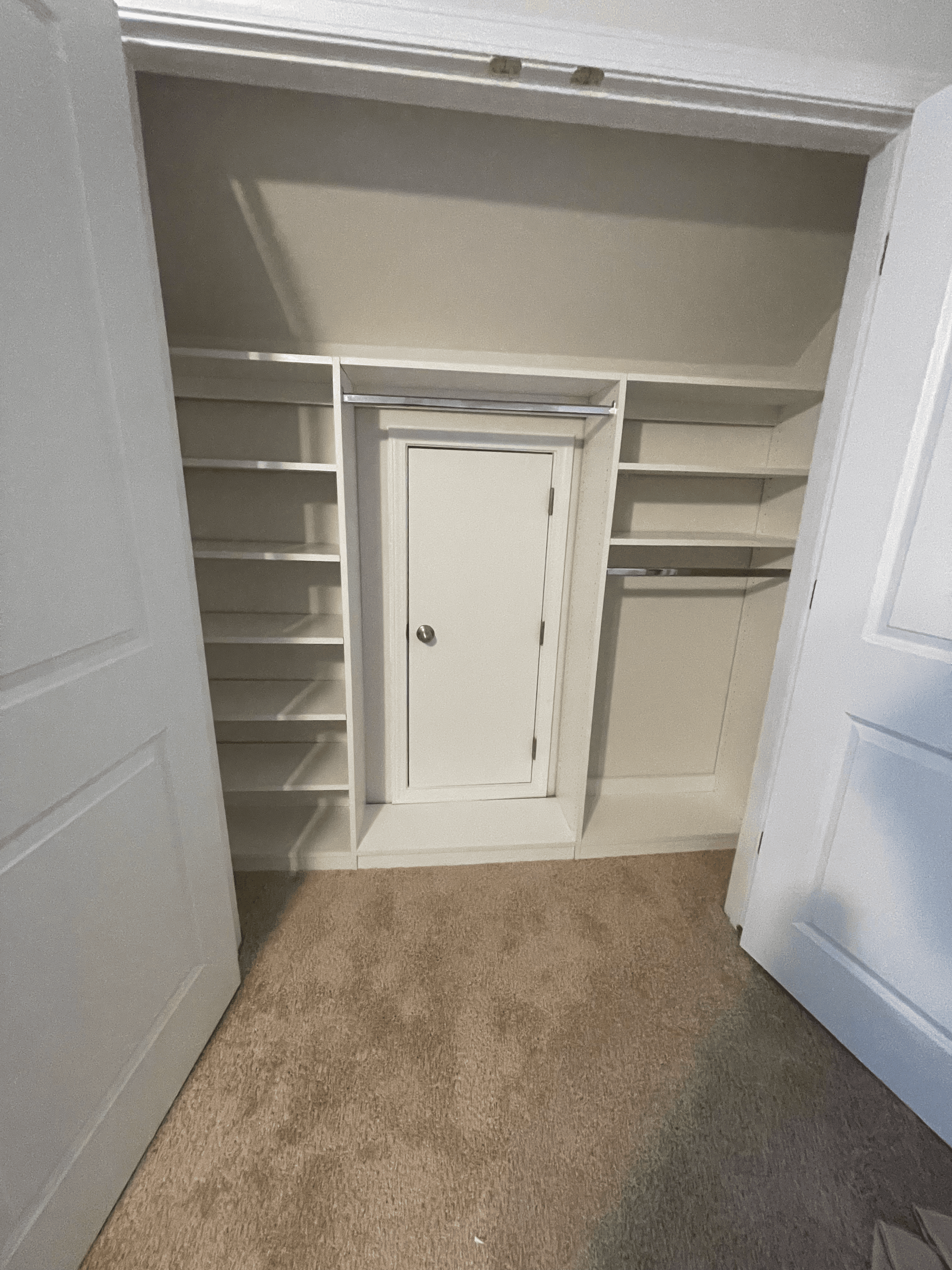 White built-in shelving units surrounding a small central access door inside a carpeted walk-in closet.